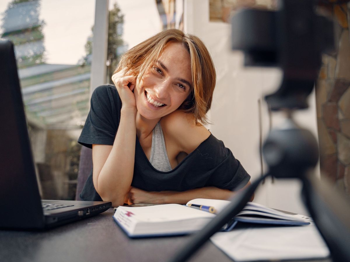 A smiling woman on a video call at her laptop, with notebooks on the desk
